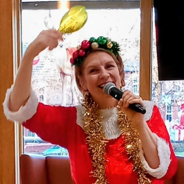 A person in a festive Santa outfit and tinsel sings into a microphone, holding up a shiny gold object during a Festive Run, with Christmas decorations and lively activities visible in the background.