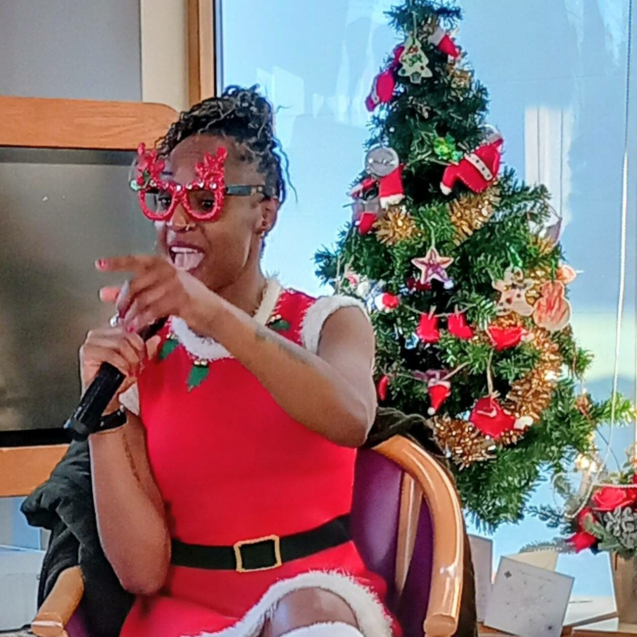 A person in festive clothing and novelty spectacles holds a microphone and points while seated before a decorated Christmas tree, ready for fun activities and music.