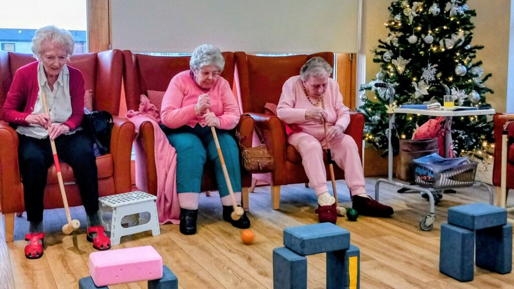 Three elderly women seated in armchairs enjoy festive activities, playing an indoor croquet game with wooden blocks as obstacles; a decorated Christmas tree stands close by.