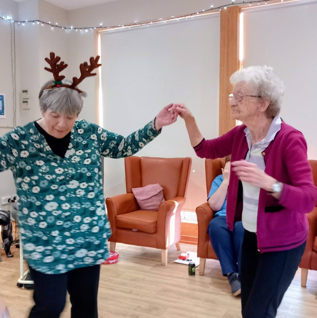 Two elderly women are dancing to music in a room; one is wearing reindeer antlers. Festive activities go on as others observe from orange chairs in the celebratory background.