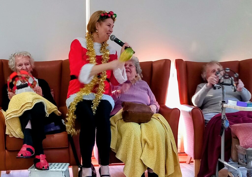 A woman dressed for a Festive Run sings into a microphone as three elderly women sit beside her, each holding a tambourine and wrapped in yellow blankets, enjoying the lively atmosphere and music.