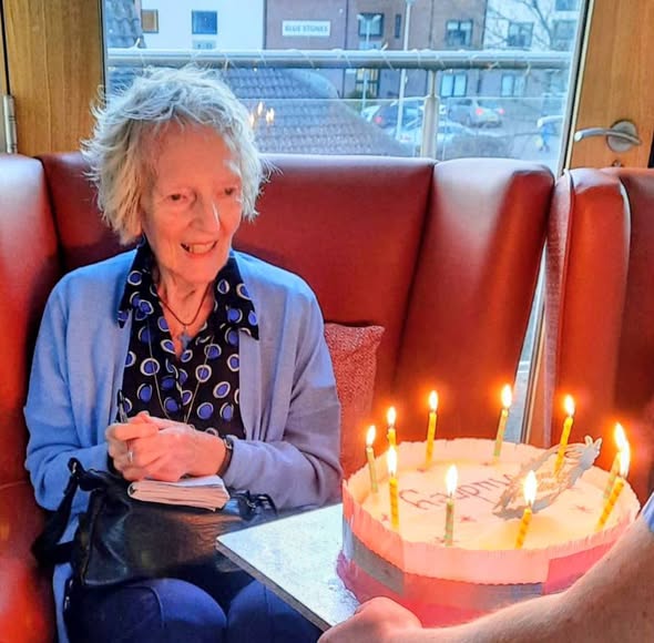 Audrey, an elderly woman, sits on a red sofa and smiles as a birthday cake with lit candles is brought to her. Happy Birthday!.