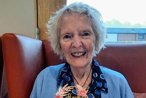 An elderly woman smiles as she holds a birthday cake adorned with candles and the name Audrey, celebrating her birthday at Abbeyfield Hoylake near West Kirby.