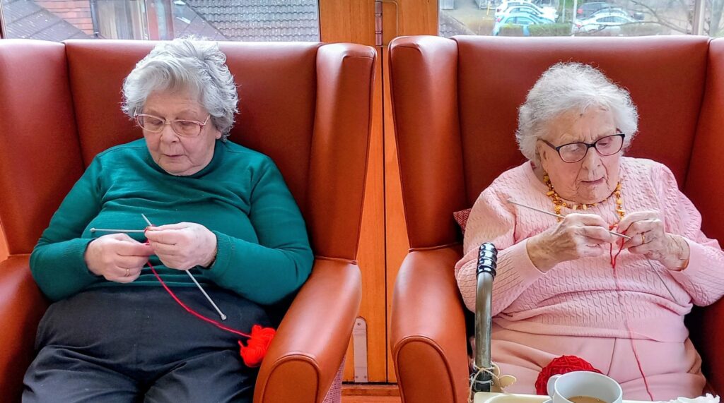 Two elderly women sit in orange armchairs by a window, knitting with red wool as they chat over drinks and balls of yarn on a tray in front of them.