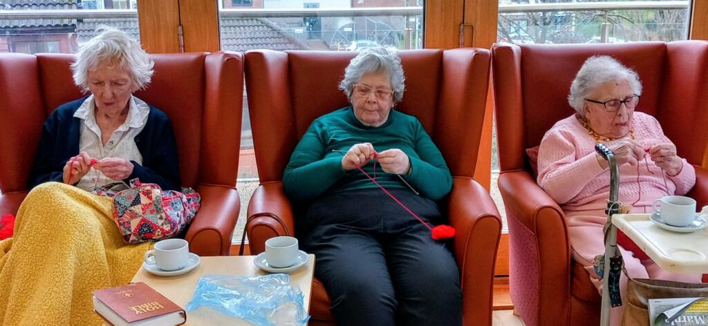 Three elderly women sit in armchairs, knitting with red wool at a Knit and Natter morning event. Cups and saucers are on the table in front of them, with a window in the background.