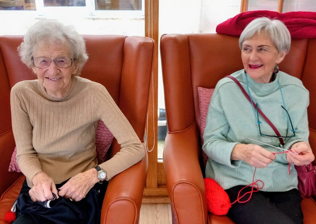Two elderly women sit in brown armchairs; one smiles at the camera, while the other enjoys a spot of social knitting with red wool, gazing to the side—capturing a cosy Knit and Natter gathering.