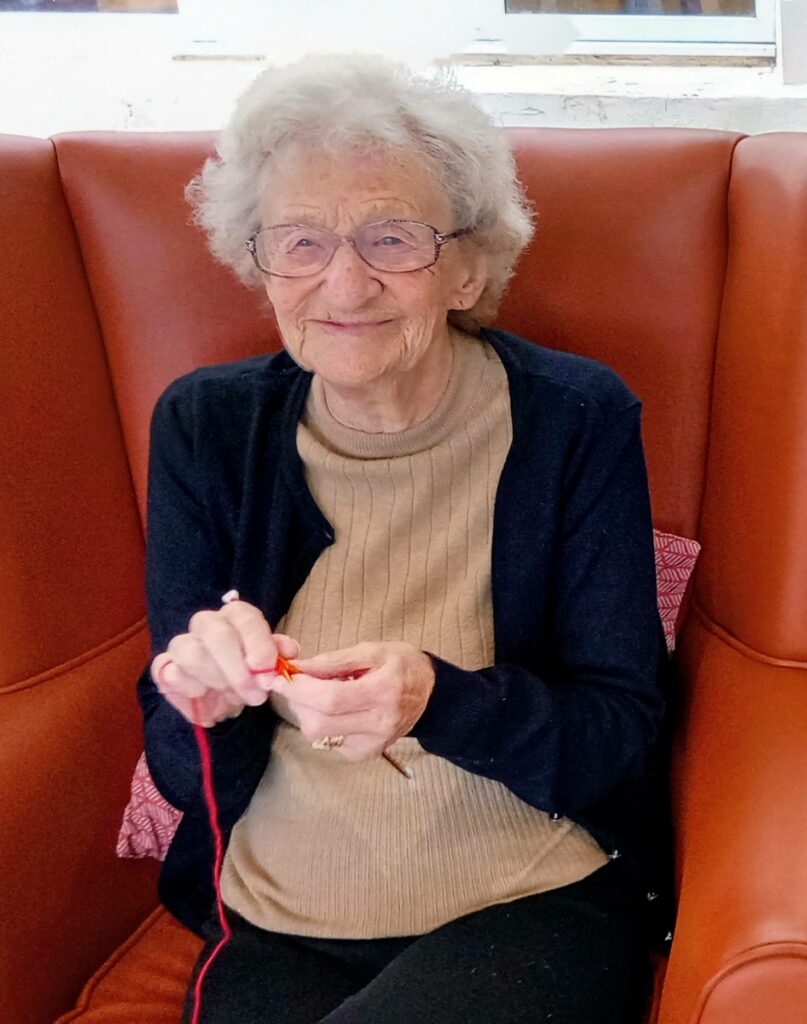 An older woman, Heather, sits in an armchair knitting, quietly enjoying her latest project before heading off to her Knit and Natter group.