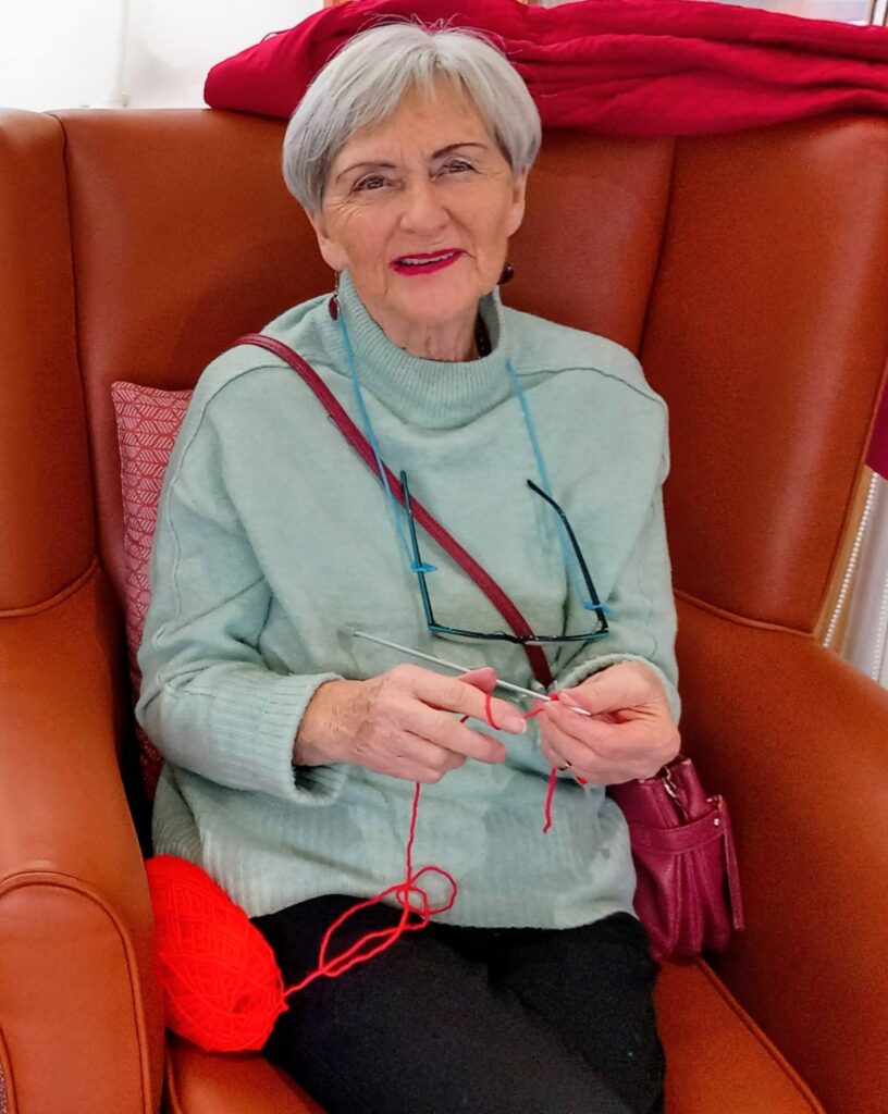 Elderly woman with short grey hair sits on a brown armchair, knitting with red wool. She wears a light green jumper in soft heather tones and has spectacles hanging on a blue cord around her neck.