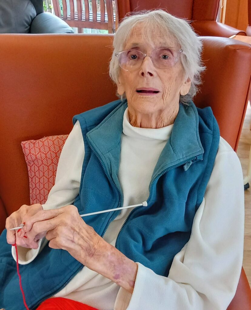 An elderly woman with glasses sits on an orange chair, holding a knitting needle and red wool, dressed for her Knit and Natter social knitting group in a white blouse and blue waistcoat.