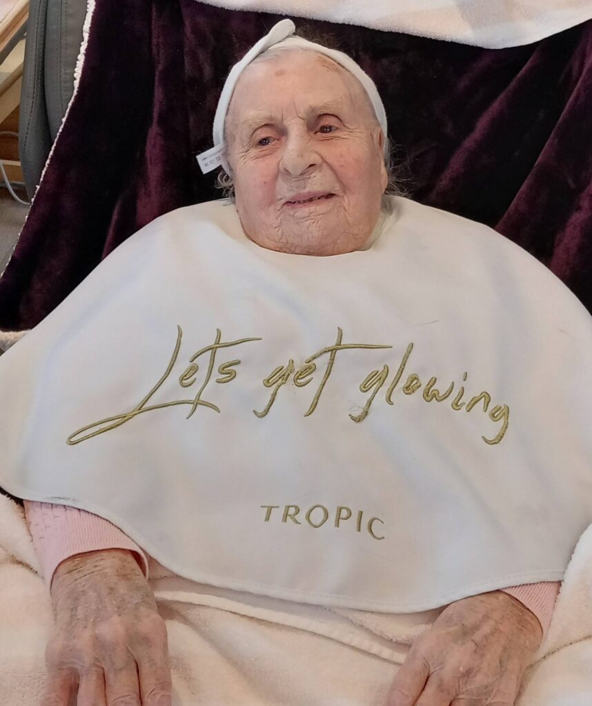 An elderly woman at Lear House sits with a white bib reading "Let's get glowing" and "TROPIC," wearing a headband and a blanket over her lap, relishing a peaceful change of pace.