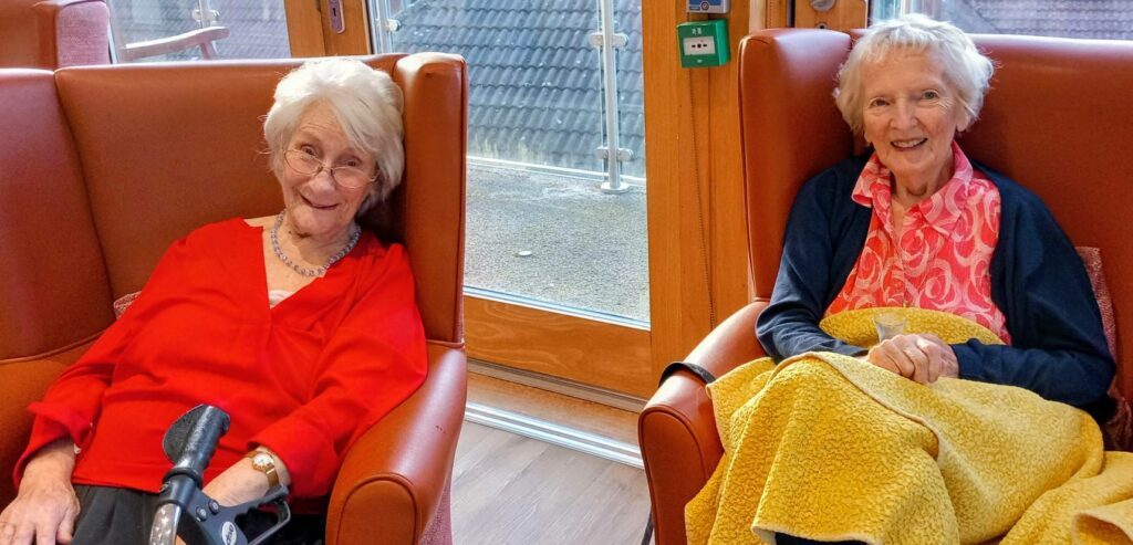 Two elderly women sit in armchairs by a window at Lear House; one has a walking frame and the other a yellow blanket on her lap. Both are smiling and enjoying a peaceful change of pace.