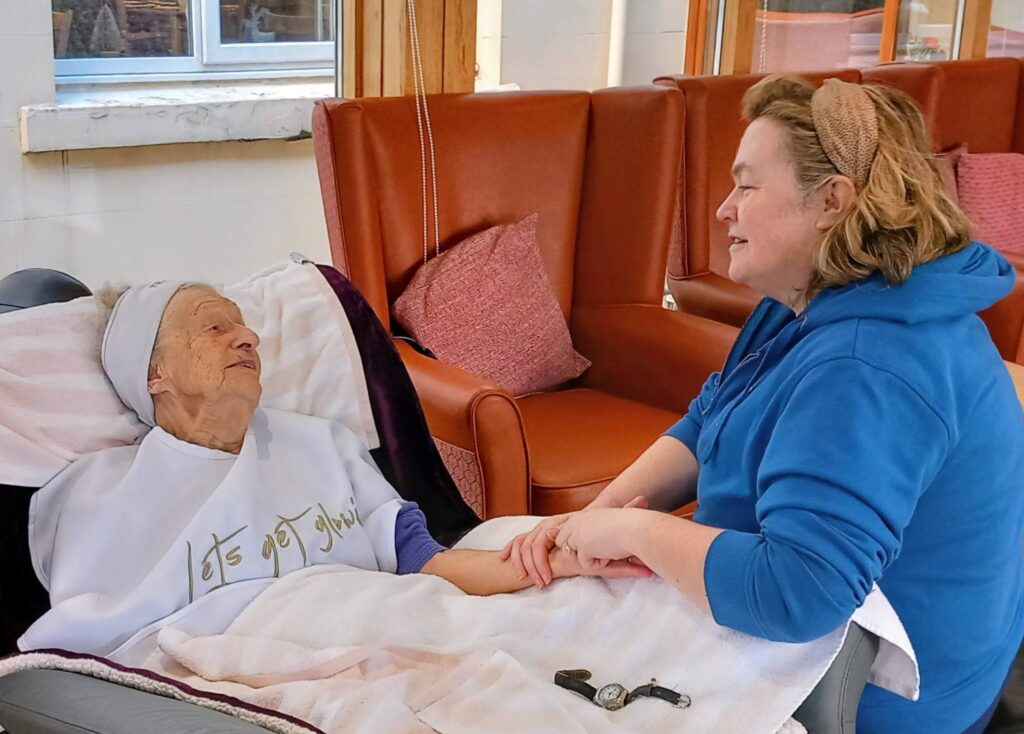 An elderly woman relaxes in a reclining chair at Lear House, holding hands with a younger woman beside her. They share a warm smile, enjoying a peaceful change of pace in the cosy indoor setting.