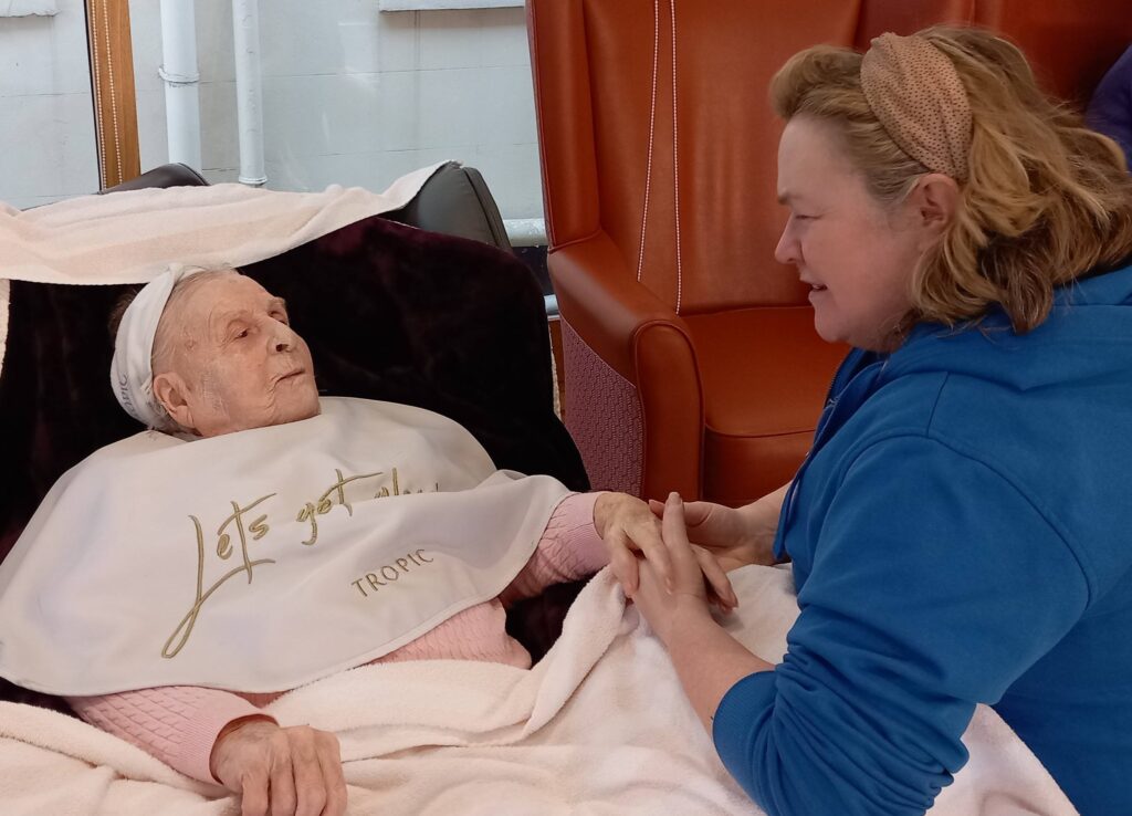 An elderly woman at Lear House lies in a chair with a blanket and bib, calmly holding hands and chatting with a younger woman in a blue jumper seated beside her, reflecting calm change.