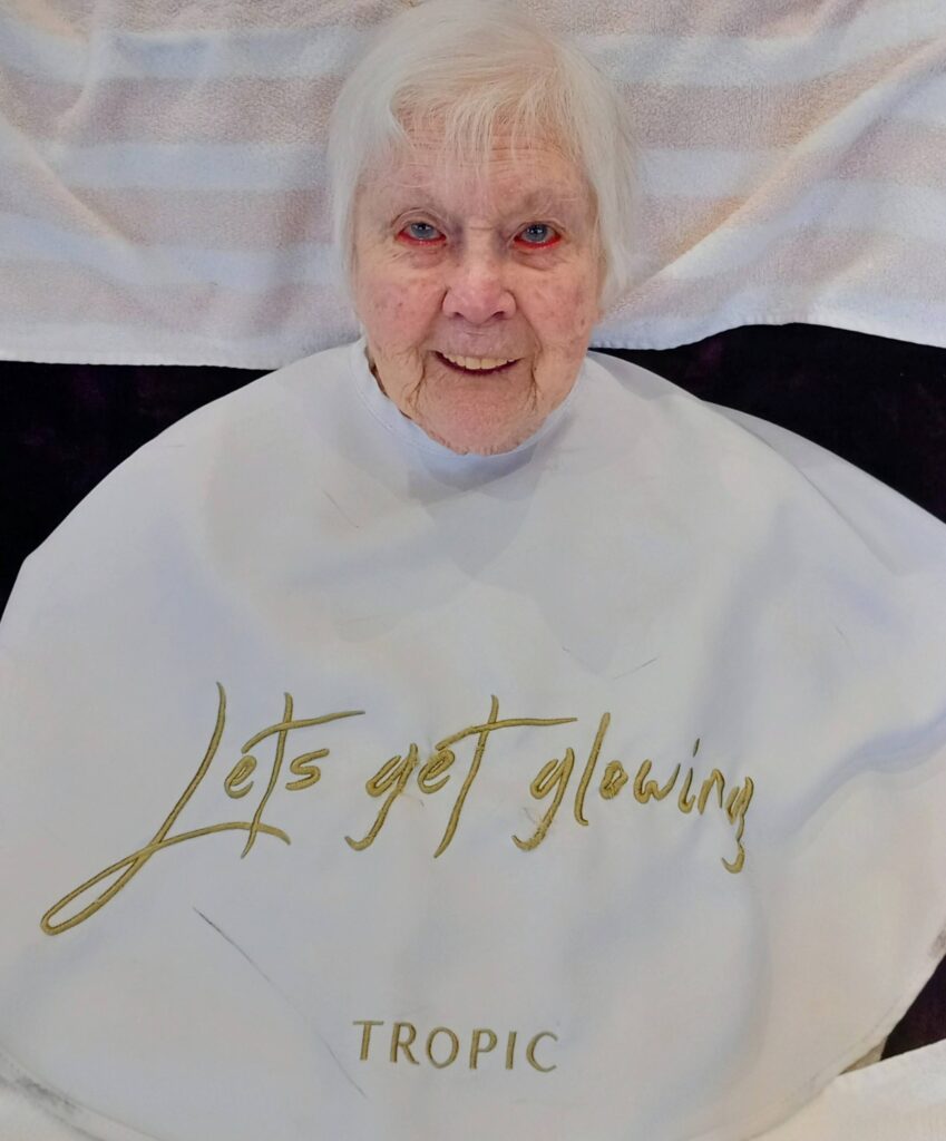 Elderly woman smiling, draped in a white cape reading “Let’s get glowing” and “Tropic,” lying on a striped towel at Lear House, enjoying a tranquil change of pace.