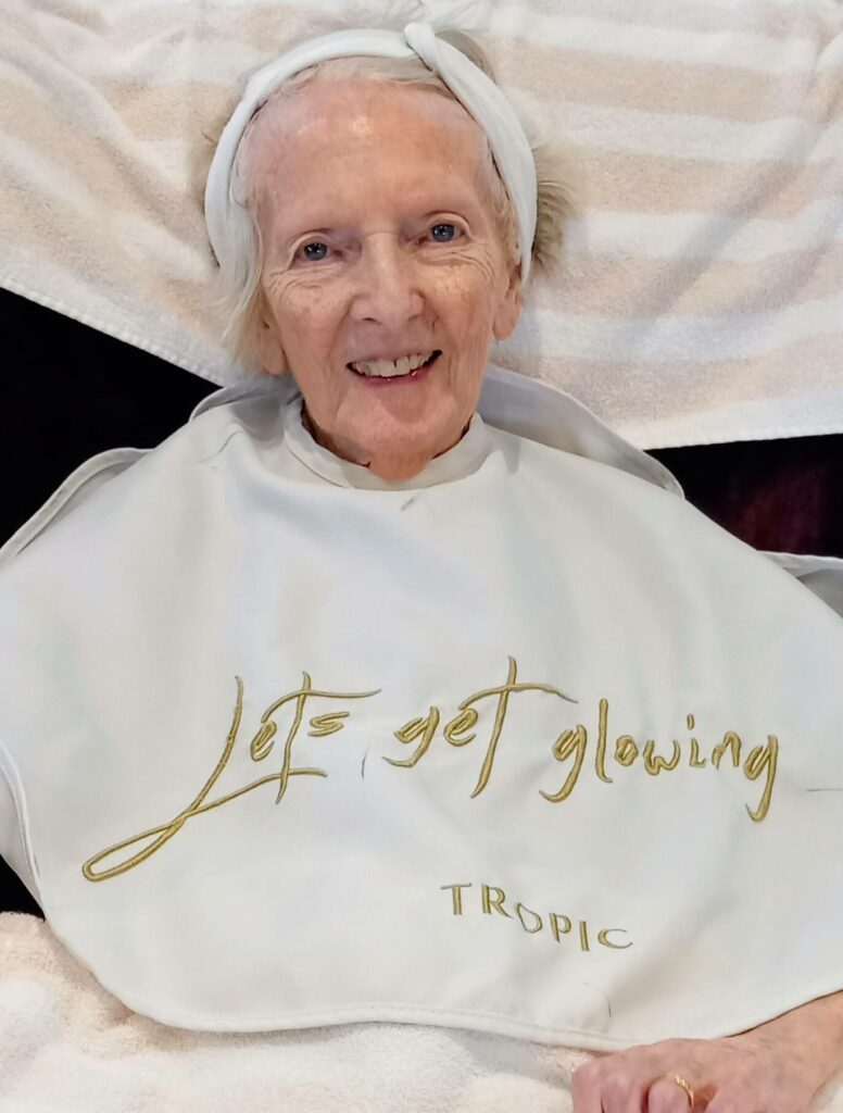 An elderly woman from Lear House smiles serenely, wearing a headband and a white cape emblazoned with "Let's Get Glowing TROPIC", seated against a backdrop of towels.