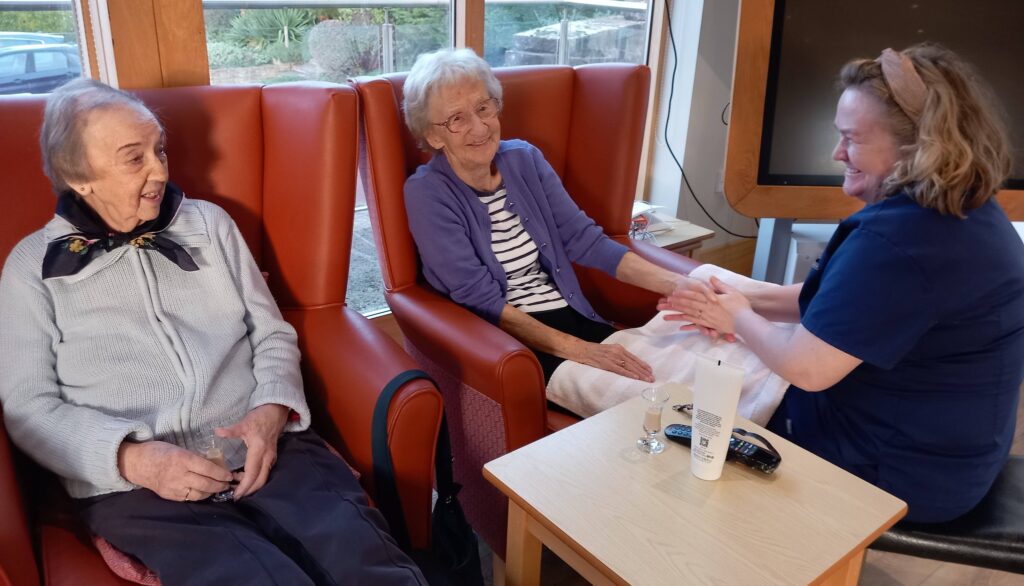 At Lear House, two elderly women sit in armchairs; one receives a hand massage from a carer while the other looks on, appreciating the tranquil change of pace. A table with lotion and spectacles is in the foreground.