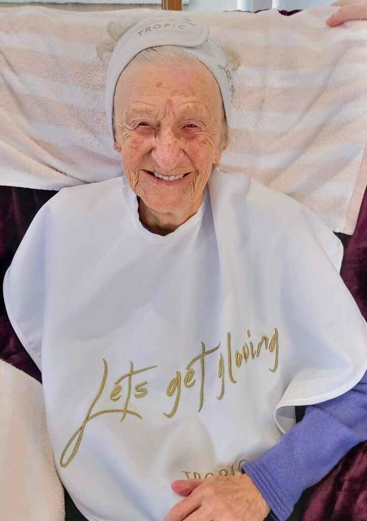 Elderly woman smiling, wearing a white headband and a "Let's get glowing" cape, enjoys a peaceful change of pace at Lear House, seated in front of a towel backdrop.