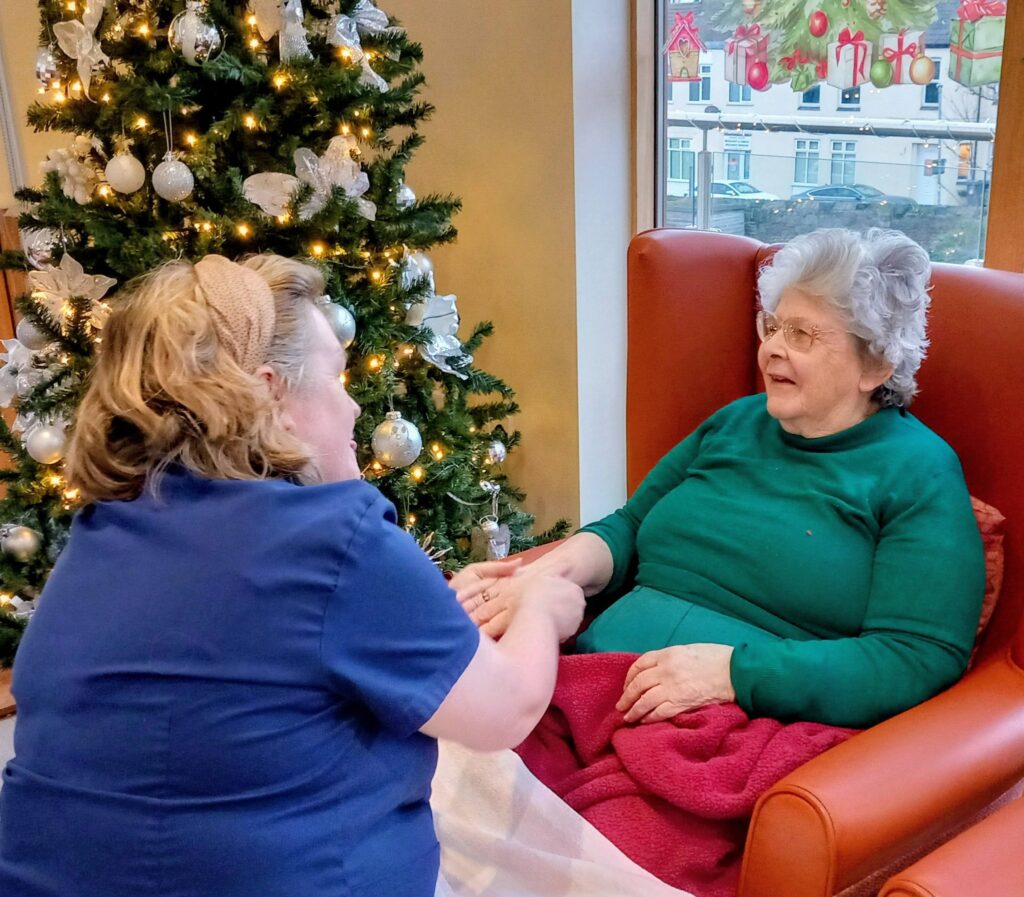A carer holds hands with an elderly woman seated in a chair near a decorated Christmas tree by a window at Lear House, creating a moment of calm change and connection.