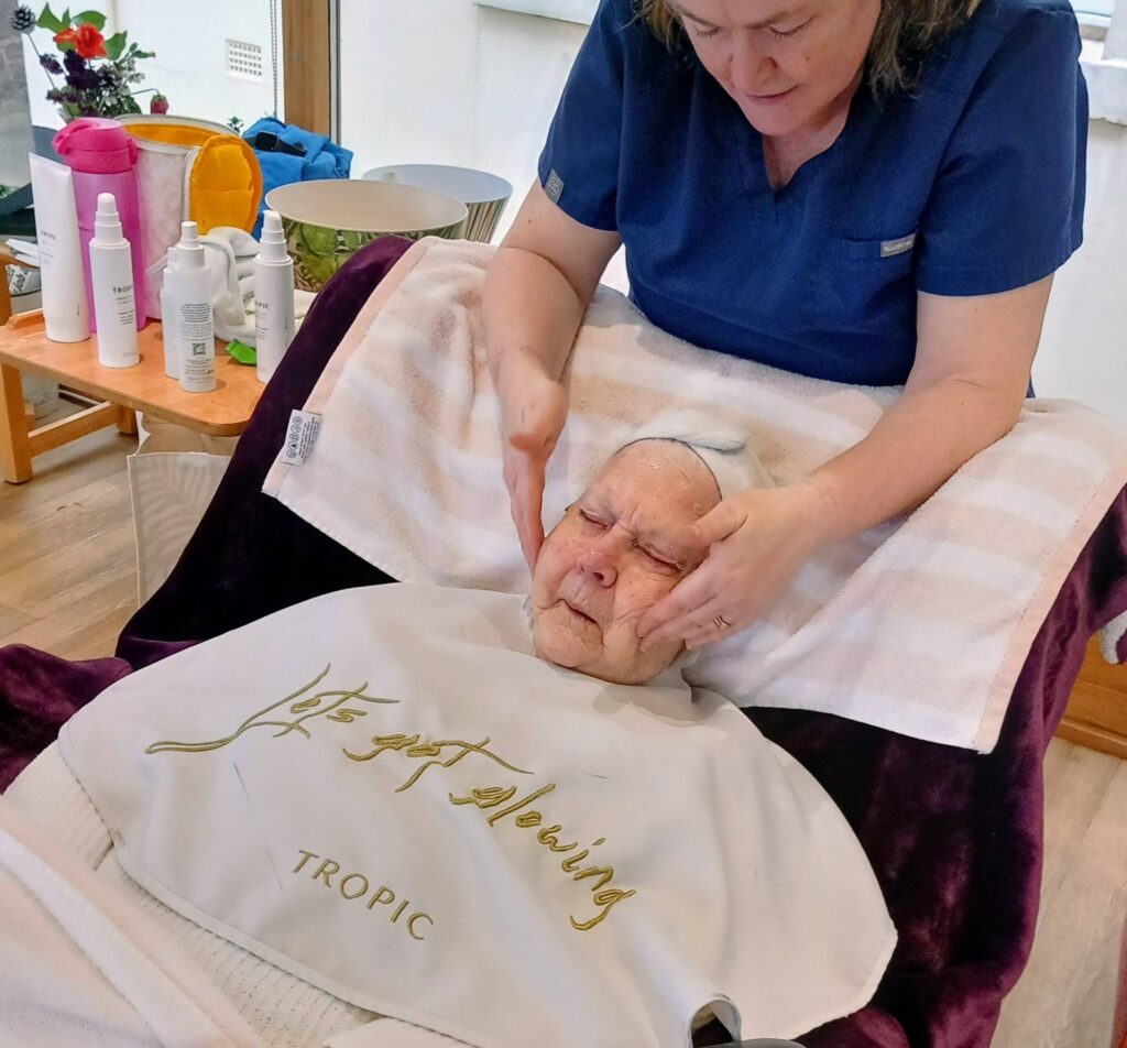 A woman in a blue uniform at Lear House gives an elderly person a facial treatment, with skincare products and towels nearby, creating a sense of calm transformation and relaxation.