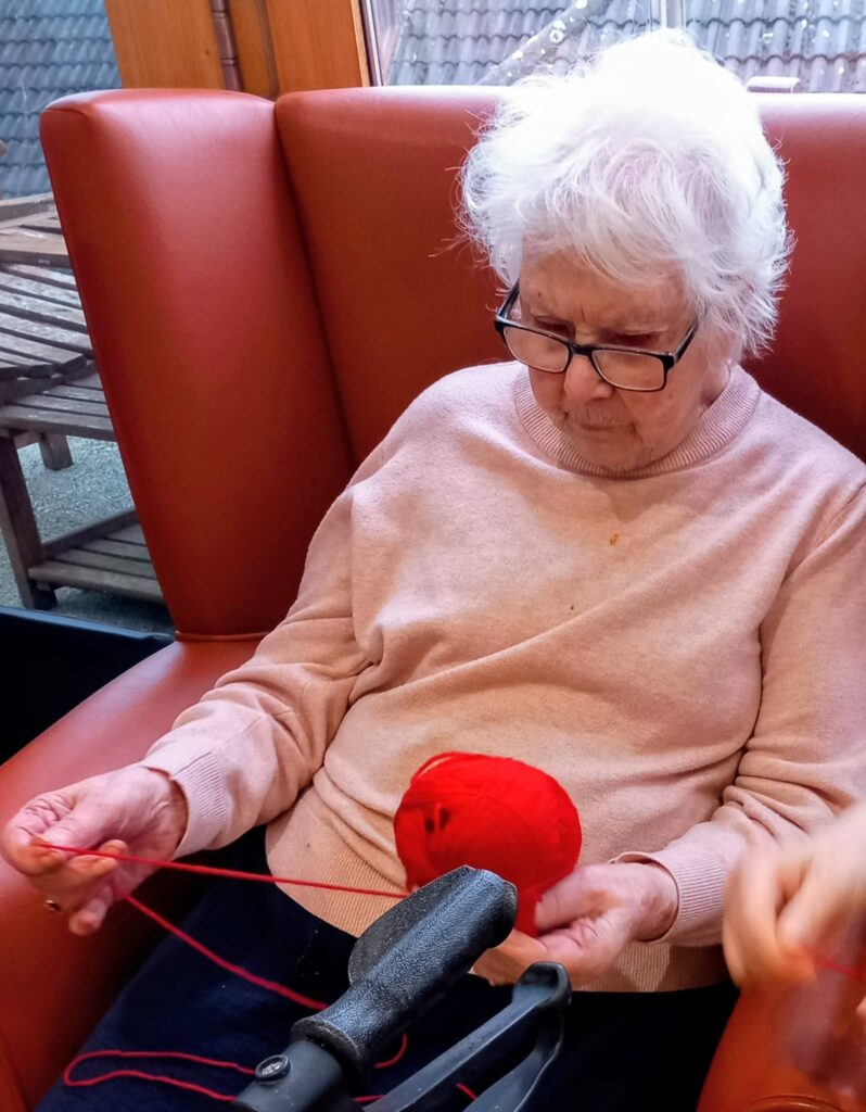 An elderly woman with white hair and glasses sits in an orange armchair, holding a ball of red wool and unwinding it during a relaxing Crafternoon.