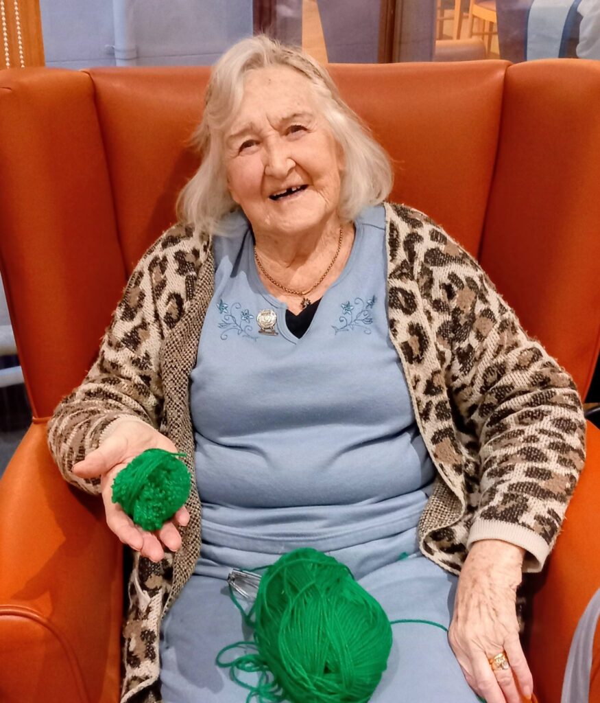 An elderly woman sits on an orange chair, smiling while holding green wool and a knitted piece—her creativity evident as she enjoys a tranquil Crafternoon in a blue dress and leopard print cardigan.