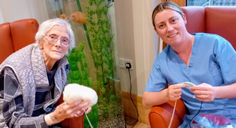 An elderly woman and a nurse sit in armchairs beside a fish tank, enjoying conversation and creativity as they knit together during a peaceful afternoon of crafting.