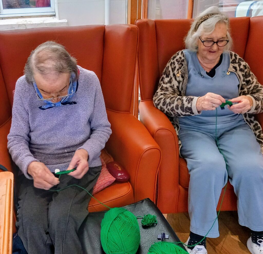 Two elderly women sit in orange armchairs, enjoying a creative Crafternoon, knitting with bright green wool. Various knitting materials are laid out on the table next to them.