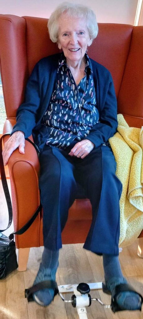 Elderly woman sits in an orange armchair, smiling, with her feet on a portable pedal exerciser and a yellow blanket next to her.