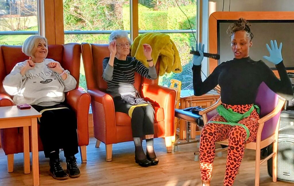 Three women sit in armchairs, following an exercise instructor wearing gloves and colourful clothing in a bright room with large windows.