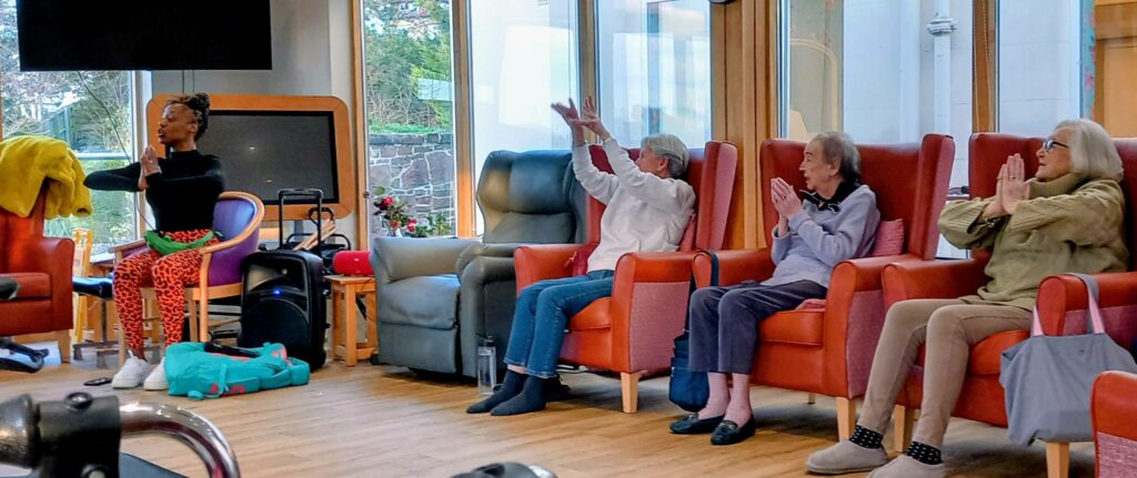 Four older adults seated in armchairs copy a fitness instructor’s movements in a bright room with large windows.
