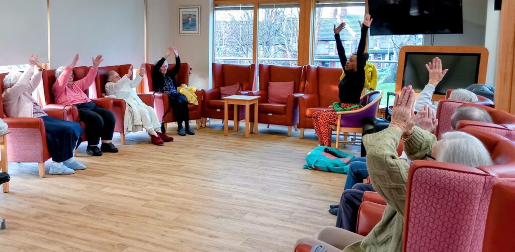 A group of elderly people sit in a circle in a brightly lit room, raising their arms as an instructor leads an exercise session.