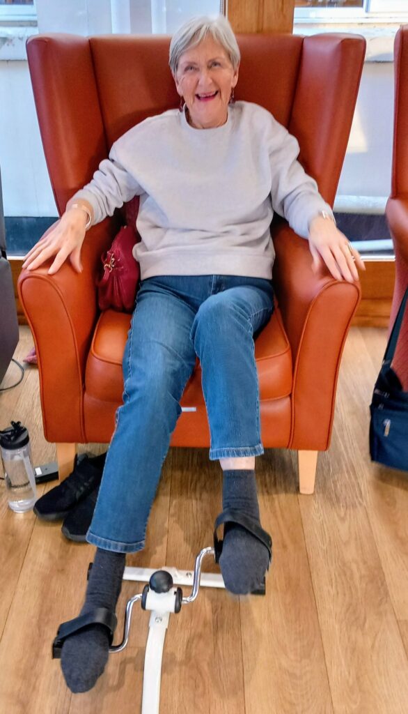 Older woman seated in an orange chair using a pedal exerciser on the floor, smiling at the camera. She is wearing a grey jumper, jeans, and dark socks.