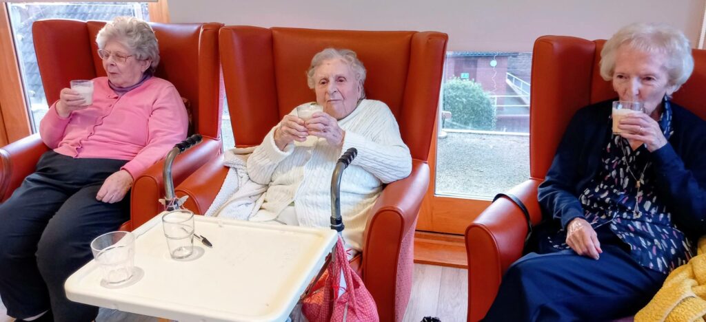 Three elderly women sit in orange armchairs by a window, each holding a glass of milk. A Zimmer frame and a tray with empty glasses are in front of them.