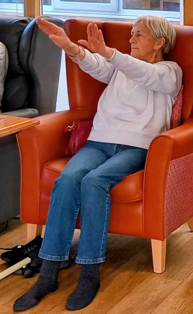 An elderly woman sits in an armchair with her arms stretched out in front of her, taking part in a seated exercise.