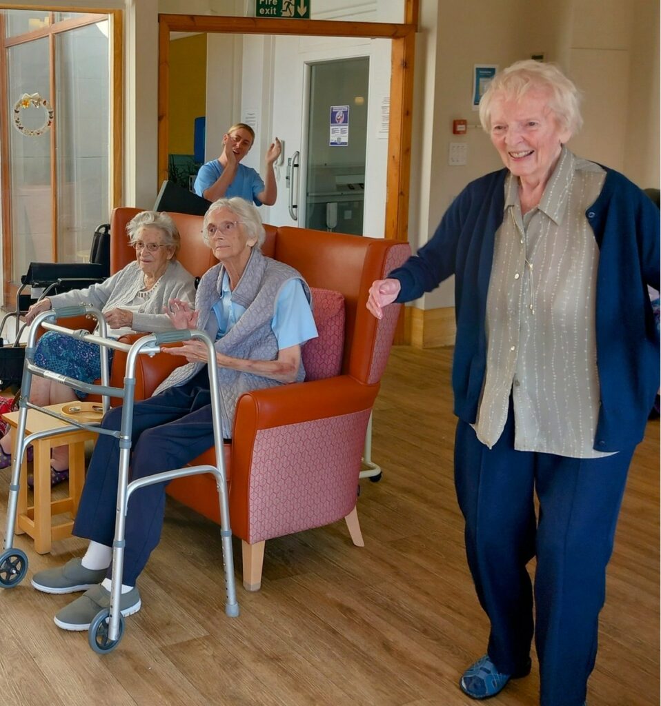 Three elderly women enjoy a pleasant afternoon in the common room; two are seated with walking frames, one stands smiling, while a staff member waves in the background.