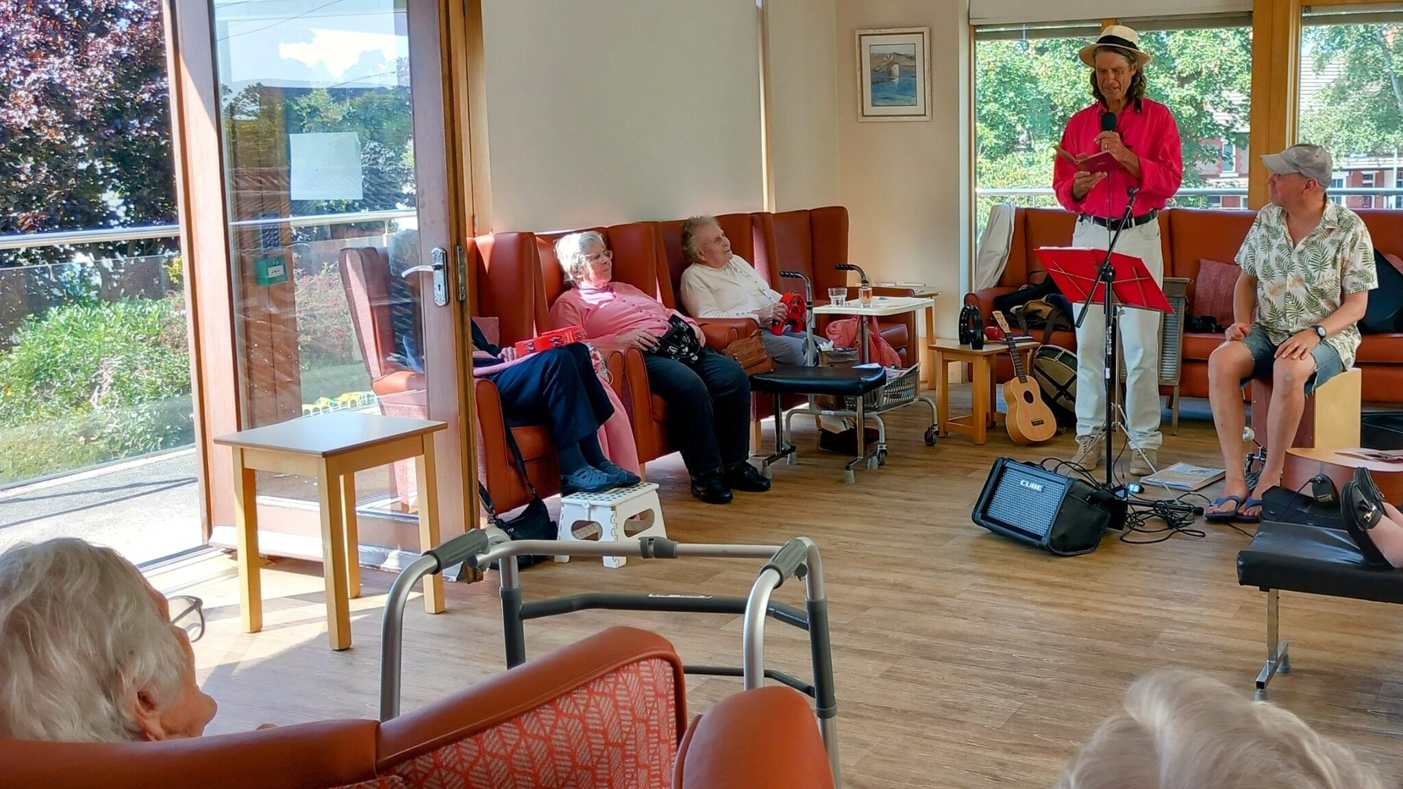 A musician stands and plays guitar for elderly people in a bright, cheerful lounge area with large windows and wooden floors on a relaxing afternoon.