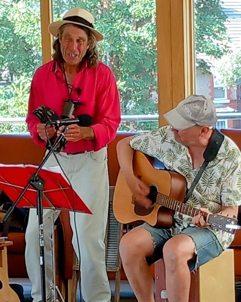 Two men perform indoors on a cheerful afternoon; one stands with a tambourine and microphone, while the other sits playing acoustic guitar. Both are casually dressed and bathed in natural light.