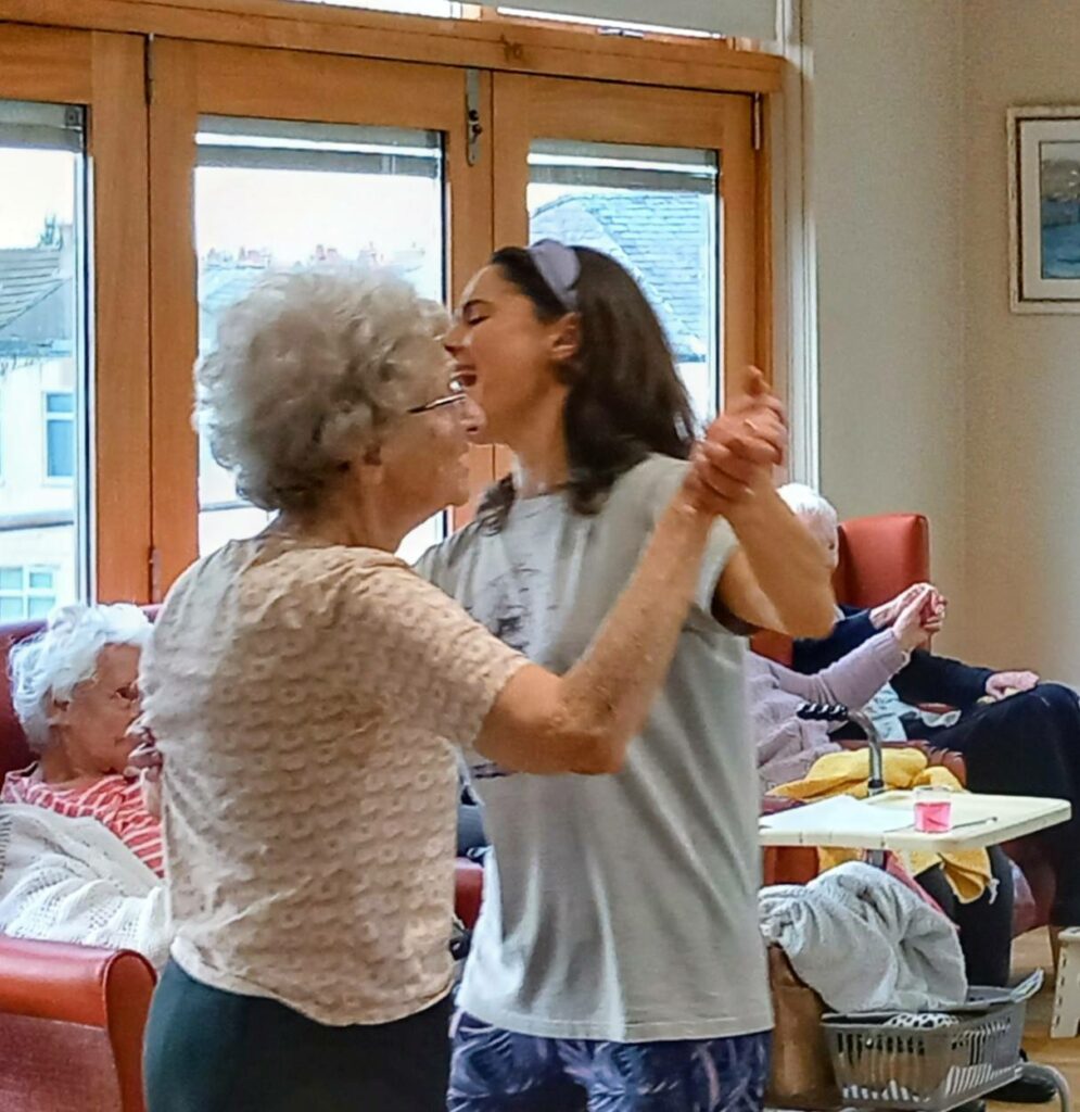 Two women, one older and one younger, are dancing together in a brightly lit room while other elderly people are seated in the background.