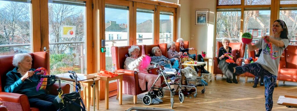 A group of elderly people sit in a sunlit room, watching a woman lead an energetic dance or exercise routine at the front. Walking frames and mobility aids are visible.