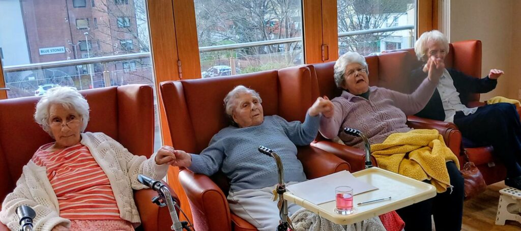 Four elderly women are seated in armchairs, holding hands. Three have blankets on their laps, and a tray with a glass sits in front of them. Large windows are visible in the background.