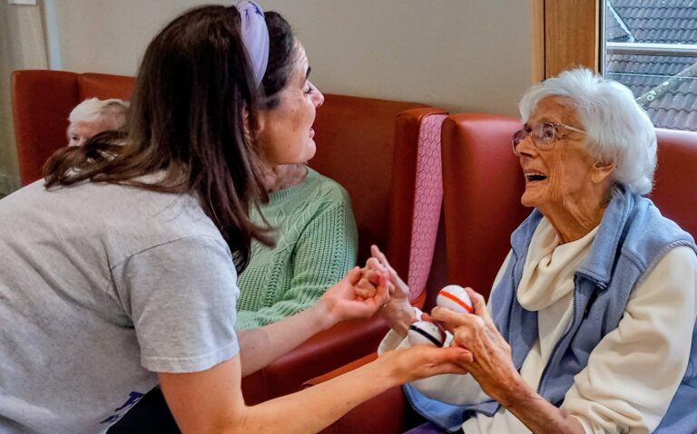 A younger woman chats with an elderly woman holding a small toy while seated in a care home; both look engaged and are smiling.