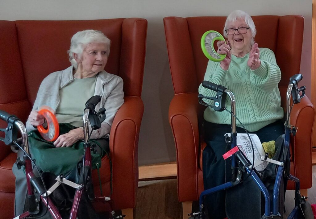 Two elderly women sit in armchairs with walking frames in front of them; one holds an orange ring, while the other smiles and lifts up green rings.