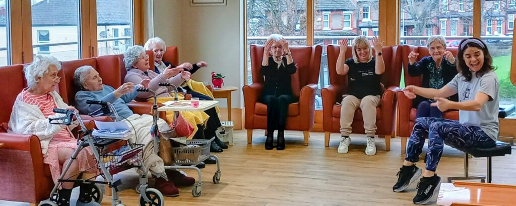A group of elderly women seated on chairs take part in a seated exercise class led by a female instructor in a bright, communal room.