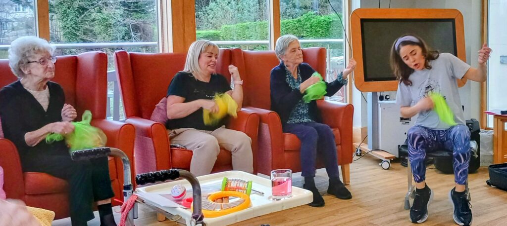 Four women sit in armchairs taking part in a group activity, waving green scarves. A younger woman is enthusiastically leading the session at the front.