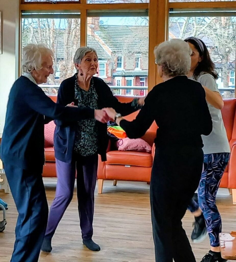 Four women, including three older adults, are holding hands and dancing together in a room with large windows and orange sofas.