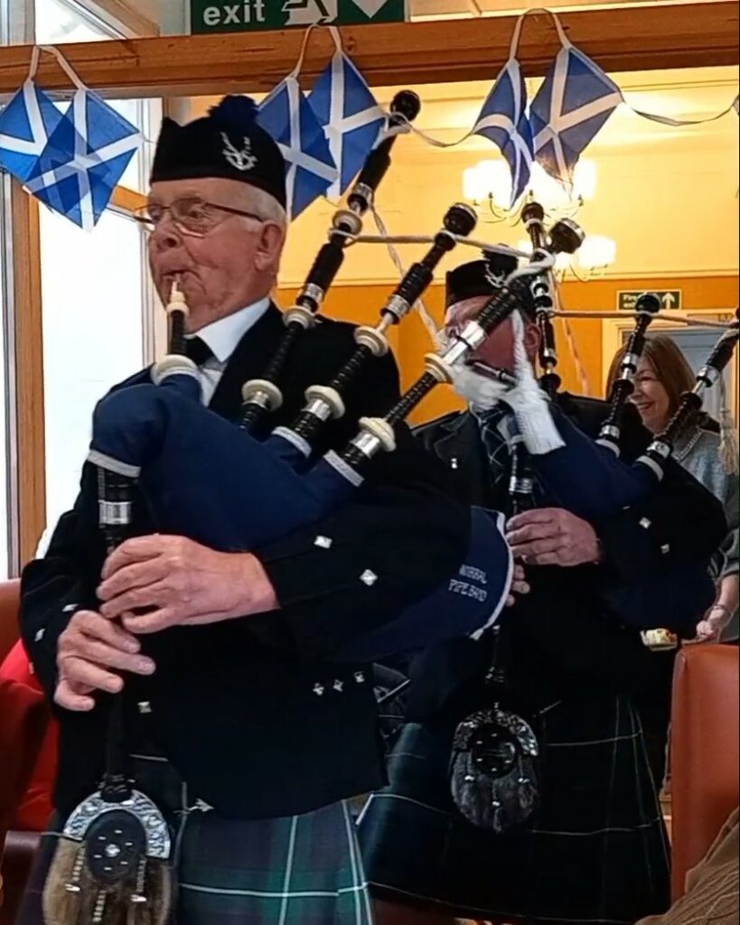 Two men dressed in traditional Scottish attire play bagpipes indoors beneath bunting featuring Scottish flags, creating a festive Burns Night atmosphere ideal for a memorable Scottish celebration.