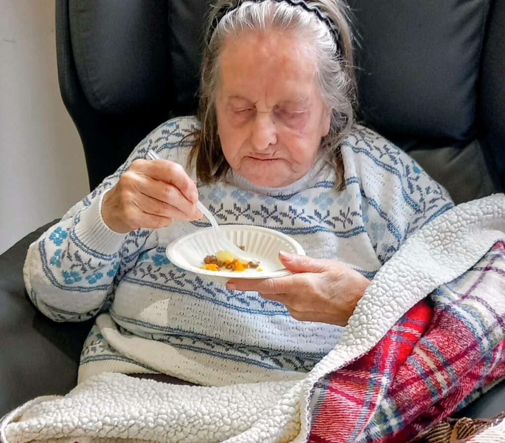 An elderly woman sits in an armchair with a blanket, holding a plate of food and eating with a fork, enjoying the warmth and tradition of a Burns Night Scottish celebration.