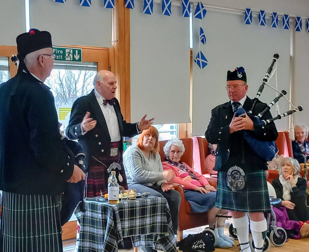 Three men in traditional Scottish dress, one playing the bagpipes, perform for a group of elderly people in a decorated room adorned with Scottish flags, capturing the Burns Night magic of an authentic Scottish celebration.