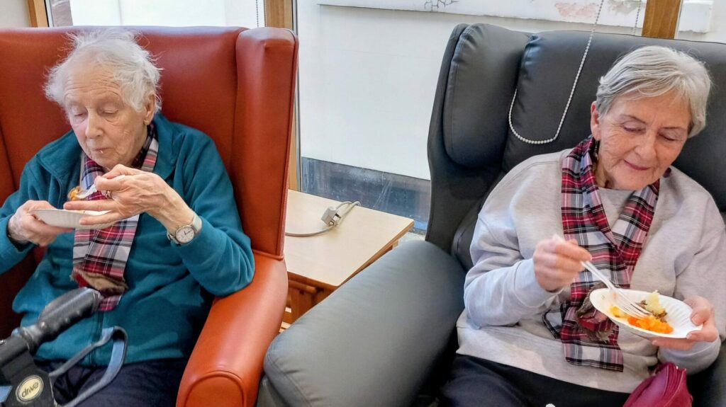 Two elderly women in tartan scarves sit in armchairs, enjoying small plates of food with plastic forks, sharing a touch of Burns Night magic during this Scottish celebration.