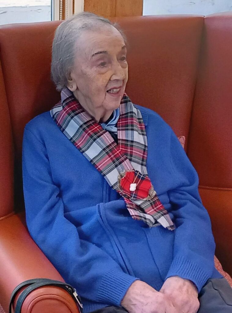 An elderly woman with short grey hair, dressed in a blue zip-up jacket and a checked scarf, sits in a red armchair and smiles, evoking the warmth of a Scottish celebration.